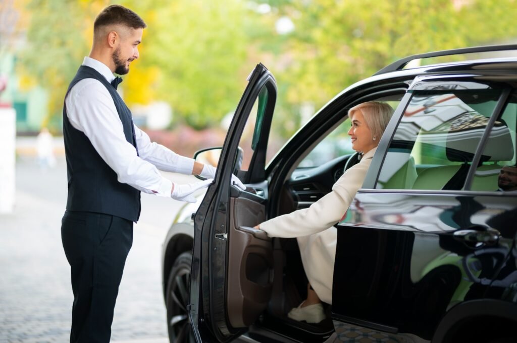 A professional chauffeur in a vest and white gloves opening the door of a black luxury SUV for a smiling female passenger in Dubai.