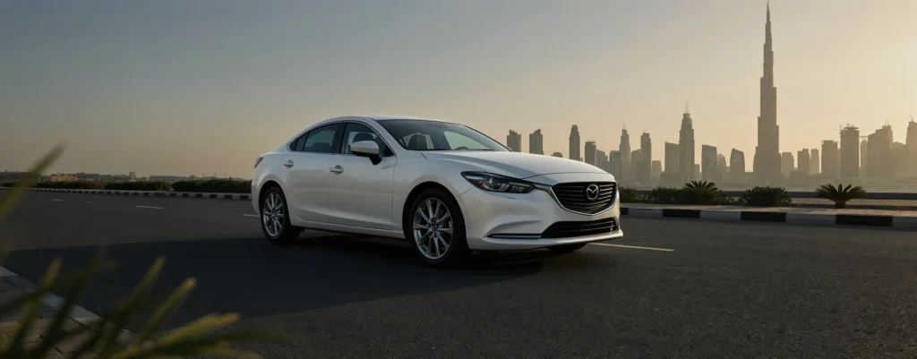 A white Mazda 6 sedan parked on an open asphalt road during the day, featuring a scenic background view of the Dubai skyline with the Burj Khalifa standing tall under a clear sky.